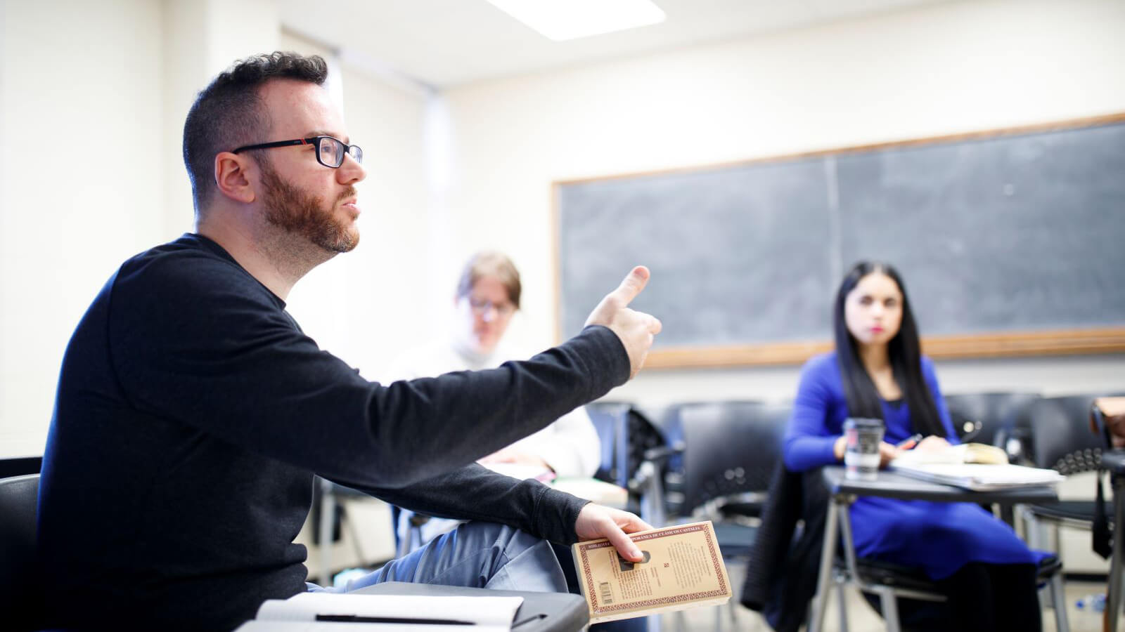 Professor and students in a classroom, seated at desks. Professor is speaking and gesturing.